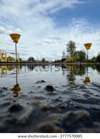 Up-close perspective of puddle on playground on blacktop with two tetherball games and playground equipment. Blue sky and clouds. Empty playground. Lonely. 