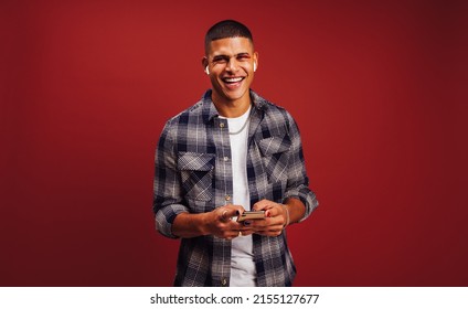 Updating my music my music playlist. Happy young man smiling cheerfully while holding a smartphone and wearing wireless earphones. Fashionable young man enjoying his favourite music in a studio. - Shutterstock ID 2155127677