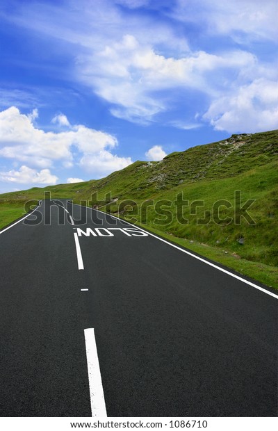 Uphill rural mountain road with a slow sign\
marking on the road, with grass verges on either side and a blue\
sky with clouds. Set in the Brecon Beacons National Park, Wales,\
United Kingdom.