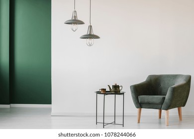 Upholstered, dark armchair and an industrial side table with a tea kettle and cup in a minimalist living room interior with a green corner - Powered by Shutterstock