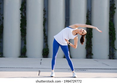 Uplifted Woman Stretching On A Concrete Paving, Doing Side Bends. Over Support Pillars. In Blue Stretchy Yoga Pants. She's Smiling At The Camera.