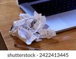 Upon a wooden table, scrunched up paper receipts lay in a pile to one side of a laptop, with a pen in the foreground.