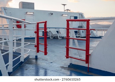 The upper deck of a white ferry features blue flooring and a red metal gate. White railings line the walkway, with deck chairs and a coastal horizon visible under a soft, early morning sky.