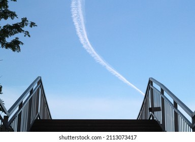 Upper edge of the stairs of the bridge with metal railings on the sides going up against blue sky. White trail from passing plane in the middle of the sky. Tree branches are visible on the left.