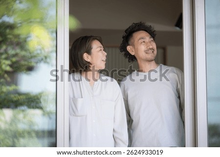 The upper half of a couple looking up and at the sky under the porch or edge of a one-story house with greenery and a view of the countryside and suburban lifestyle.