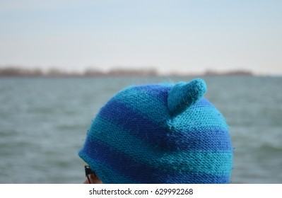 Upper part of a blue winter hat with ears on the head of a young boy in front of a waterfront background.