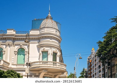 Upper part of corner white building facade with dome, spire, arched windows, round balcony with balustrade against city buildings, blue sky.