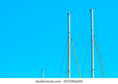 Upper parts of sailboat masts against a clear blue sky. Masts are equipped with various rigging lines and small devices at their tops, creating a visually scene due to the contrast with vibrant sky - Powered by Shutterstock