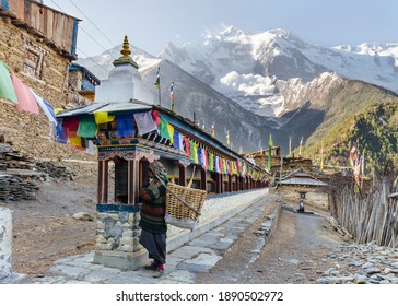 Upper Pisang, Nepal - 03.01.2017: Old local woman spinning prayer wheels along a Mani wall with a snow avalanche on Annapurna II mountain in the background