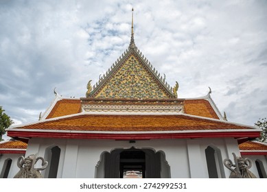 The upper portion of a white temple building at Wat Arun, in Bangkok, Thailand. The prominent front gable is elaborately decorated with green and gold mosaic patterns and topped with a golden spire.
