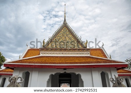 The upper portion of a white temple building at Wat Arun, in Bangkok, Thailand. The prominent front gable is elaborately decorated with green and gold mosaic patterns and topped with a golden spire.