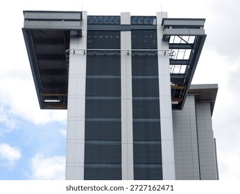 The upper section of a modern skyscraper featuring a unique cantilevered design and a stark industrial aesthetic. The building is characterized by its vertical grey panels and dark horizontal louvers, creating a strong sense of geometric precision against the bright sky. On either side, massive rectangular platforms extend outward, revealing the intricate structural steelwork and mechanical elements underneath. The overall composition highlights a fusion of contemporary engineering and minimalist urban design, emphasizing the scale and functionality of the high-rise structure.