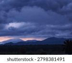 The upper three-quarters of the frame is dominated by an expansive, dramatic sky filled with thick, churning storm clouds. The clouds are cast in deep indigo, slate grey, and dark blue tones, creating a heavy and moody atmosphere. Beneath the dense cloud cover, a soft, narrow band of pastel pink and pale orange twilight breaks through right along the horizon, contrasting sharply with the dark clouds above. In the mid-ground, a series of rolling mountain ranges is visible in silhouette. The peak on the left is partially shrouded in soft fog or mist, while a faint vertical antenna tower stands on the ridge line in the center.