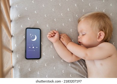 Upper view of baby napping in his bed next to phone playing lullaby in app for baby sleep with child tracker function and parents remote control settings. Maternity and technology. Healthy sleep - Powered by Shutterstock