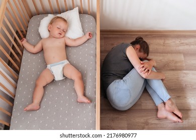 Upper view of cute baby napping in his bed next to his mother lying on floor crying, feeling desperate and lonely, suffering postnatal depression. Difficulties of maternity