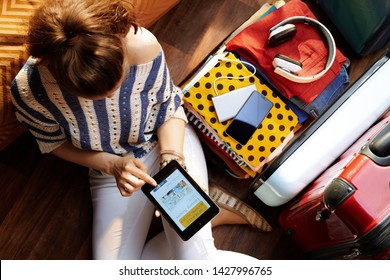 Upper view of elegant woman in white pants and striped blouse at modern home in sunny summer day using mobile app hotel booking near open travel suitcase.