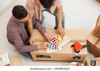 Upper View Of Two Unrecognizable Men Sitting On The Floor And Looking At Color Pattern Samples, On A Cardboard Box Of A Moving. Moving Into A New Home Concept, Renovation, Repairing, Designing.