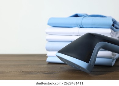 Upright steamer and stack of folded shirts on wooden table near white wall indoors, closeup. Space for text