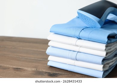 Upright steamer and stack of folded shirts on wooden table near white wall indoors, closeup. Space for text