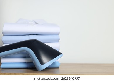 Upright steamer and stack of folded shirts on wooden table near white wall indoors, closeup. Space for text