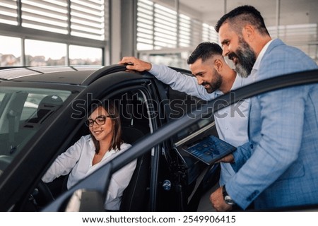 In an upscale car showroom, two knowledgeable salesmen eagerly demonstrate the advanced functionalities of a sleek new car to an interested woman seated in the driver’s seat.
