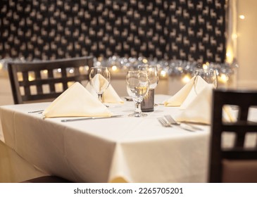 Upscale restaurant dining room table with place settings, napkins, wooden chairs, white tablecloth, candle light and holiday background with lights.  - Shutterstock ID 2265705201