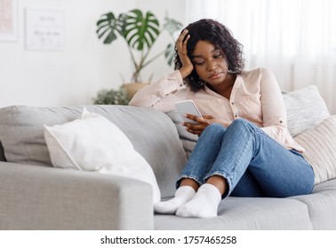 Upset african american girl using smartphone while sitting on couch at home, having depression, waiting for importrant call, free space