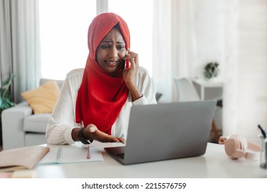 Upset African American Woman In Hijab Looking At Laptop Screen And Talking On Phone, Having Problems With Her Job, Sitting At Desk In Living Room Interior, Copy Space