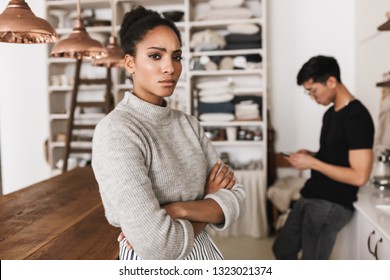 Upset African American Woman Holding Hands Together Sadly Looking In Camera With Asian Man Holding Cellphone On Background. Young International Couple In Quarrel Spending Time On Kitchen At Home