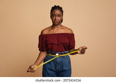 Upset african american young woman in casual outfit measuring her waist over beige studio background, unhappy with results of healthy diet or sporty lifestyle black lady, copy space