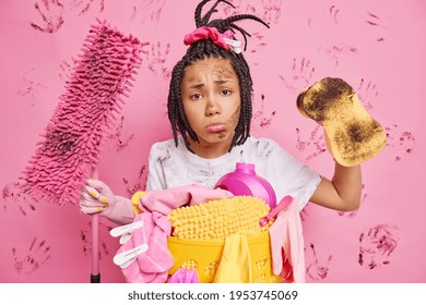 Upset Afro American woman poses near laundry basket with dirty linen holds dirty mop and sponge uses cleaning equipment brings house in order looks sadly at camera has dirty face. Domestic chores - Powered by Shutterstock