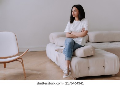 Upset Afro Brazilian Young Woman Sitting On Cozy Sofa At Home Alone. Frustrated African American Girl In White T- Shirt, Blue Jeans At Home Disappointed By Financial Troubles, Divorce, Rent Price.