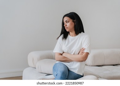 Upset Afro Brazilian Young Woman Sitting On Cozy Sofa At Home Alone. Frustrated African American Girl In White T- Shirt, Blue Jeans At Home Disappointed By Financial Troubles, Divorce, Rent Price.