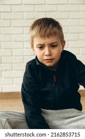 Upset, Angry Boy Of 6-7 Years Old Sits Near The Wall. Portrait Of A Boy, Front View