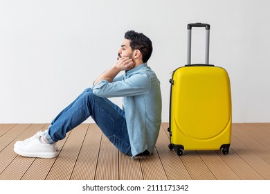 Upset Arab Male Tourist Sitting Near Suitcase And Looking Away At Copy Space, Waiting For Flight Over Light Wall. Man Traveling Abroad On Weekends Getaway. Air Flight Journey Concept