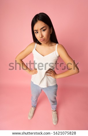 Upset asian girl looking at camera, holding hands on hips over pink background, high angle view