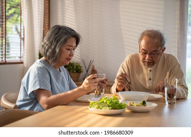 Upset Asian Senior Elderly Retired Grandparent Stay Home With Painful Face Sit On Eating Table In House. Depressed Mature Older Man And Woman Couple Feeling Unhappy, Lonely And Missing Family At Home.