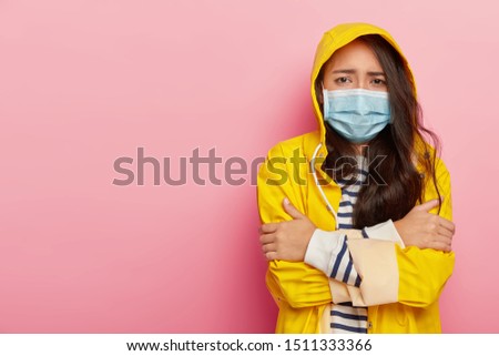 Upset Asian woman trembles from cold, has virus transmitted through airbone droplets, wears protective medical mask, yellow raincoat with hood, stands against pink wall, has contagious disease