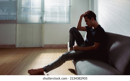 An upset Asian young man sitting alone near a window on a couch at home office sad and looking down, having problems. Copy space. Loneliness, depression, suffering, financial hangover concept ideas. - Powered by Shutterstock