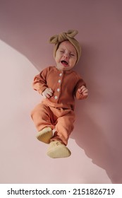 Upset baby in pajama crying. Top view of sad baby in warm sleepwear and trendy headband crying loudly while lying on pink background