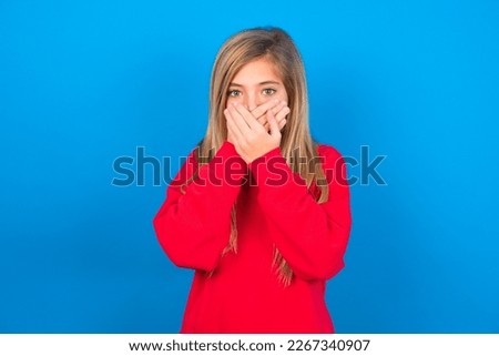 Upset Beautiful caucasian teen girl wearing red sweatshirt over blue background, covering her mouth with both palms to prevent screaming sound, after seeing or hearing something bad.