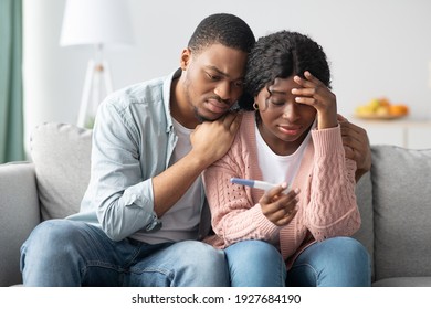 Upset black couple with negative pregnancy test sitting on couch at home, hugging and looking at test, free space. Disappointed african american man and woman suffering from infertility problem