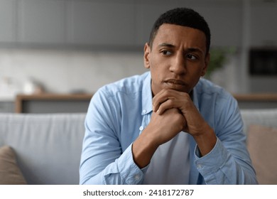 Upset black man having problems, sitting on couch at home and thinking, having heavy thoughts, break up with lover or financial difficulties