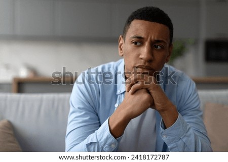 Upset black man having problems, sitting on couch at home and thinking, having heavy thoughts, break up with lover or financial difficulties