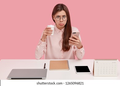 Upset brunette female office perfectionist has unhappy expression, drinks takeaway coffee, holds mobile phone, wears round spectacles, elegant shirt, likes everything is in proper order. Perfectionsim