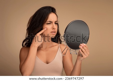 Upset brunette young woman wearing beige top looking at mirror and touching her face over beige studio background, suffering from skin problems. Acne, wrinkles, dull skin, early premature aging