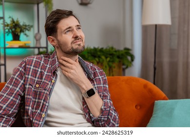 Upset Caucasian ill sick man holds hands over sore throat. Painful neck and frowning, thyroid disorders, suffering sore throat, tonsils inflammation. Young guy sitting on sofa at home room apartments - Powered by Shutterstock