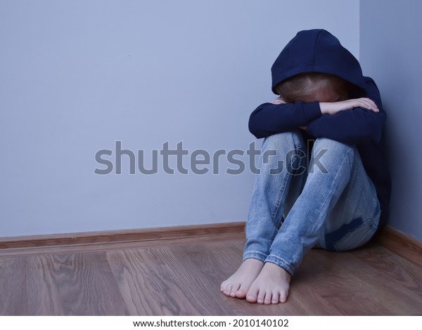 An upset child or teenager sits on the floor in the\
corner with a hood pulled over his head, hugging his knees and\
hiding his face. Parenting problems, social problems,\
misunderstanding of parents.\
