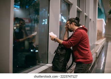 A upset, confused young man with tickets in his hands at the airport gate at night. A sad guy, a tourist with a backpack, hand luggage, looks sullenly at the ticket. Delay, cancellation of the flight - Shutterstock ID 2223747127