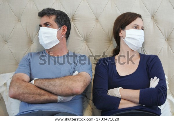 Upset\
couple in self isolation wearing face mask and protective gloves\
bored in home bedroom sitting on bed looking\
away.